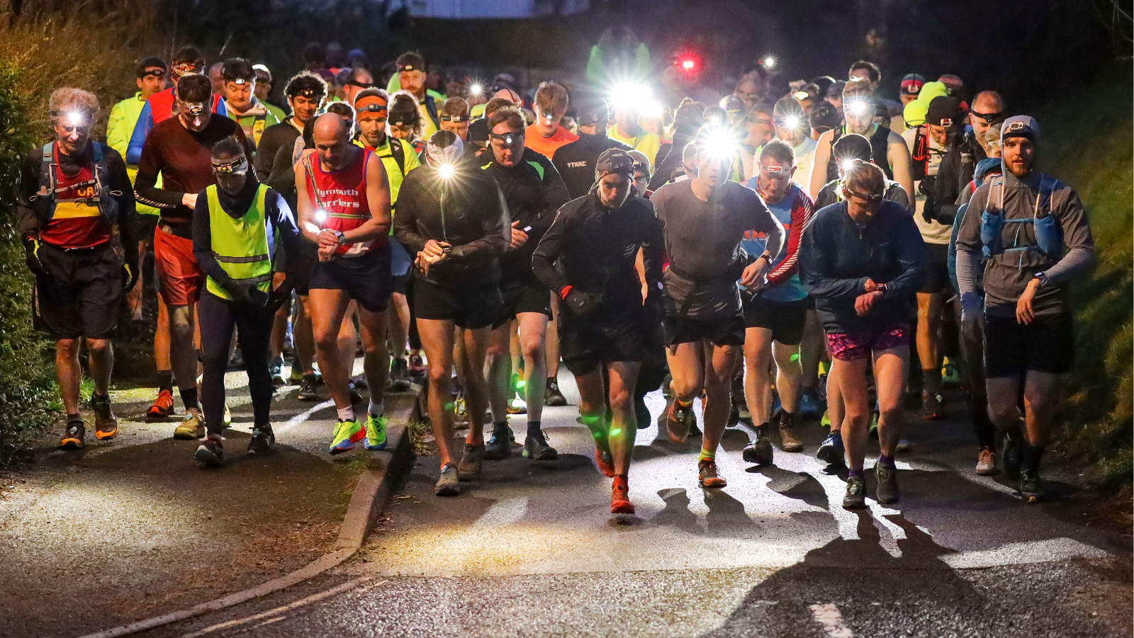 A group of runners, wearing headlamps and reflective gear, start a race on a paved road at night. The crowd includes men and women, all concentrated and determined, with others visible in the background. The road is dimly lit by their headlamps.