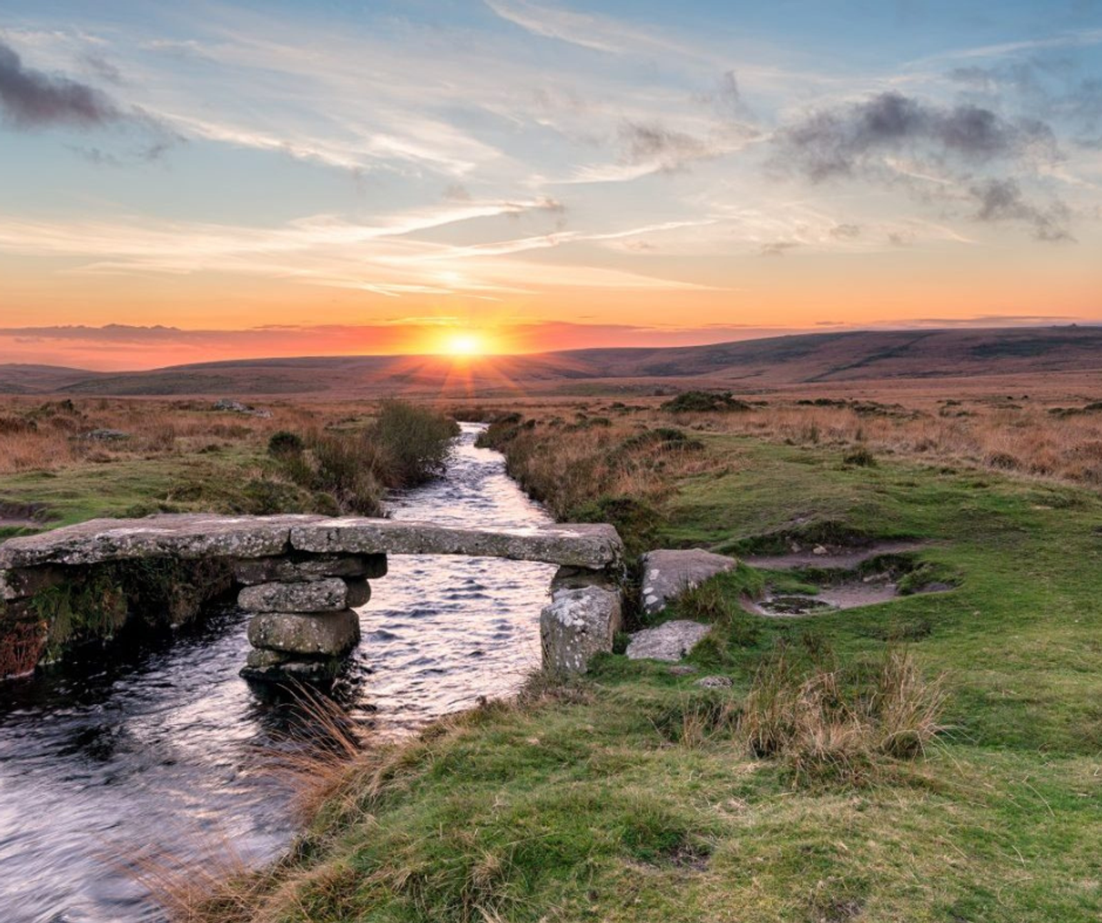 A picturesque sunset over a tranquil stream with a rustic stone footbridge in an expansive open moorland. The sky is painted in hues of orange, pink, and blue as the sun descends near the horizon, with grassy fields stretching in the distance.