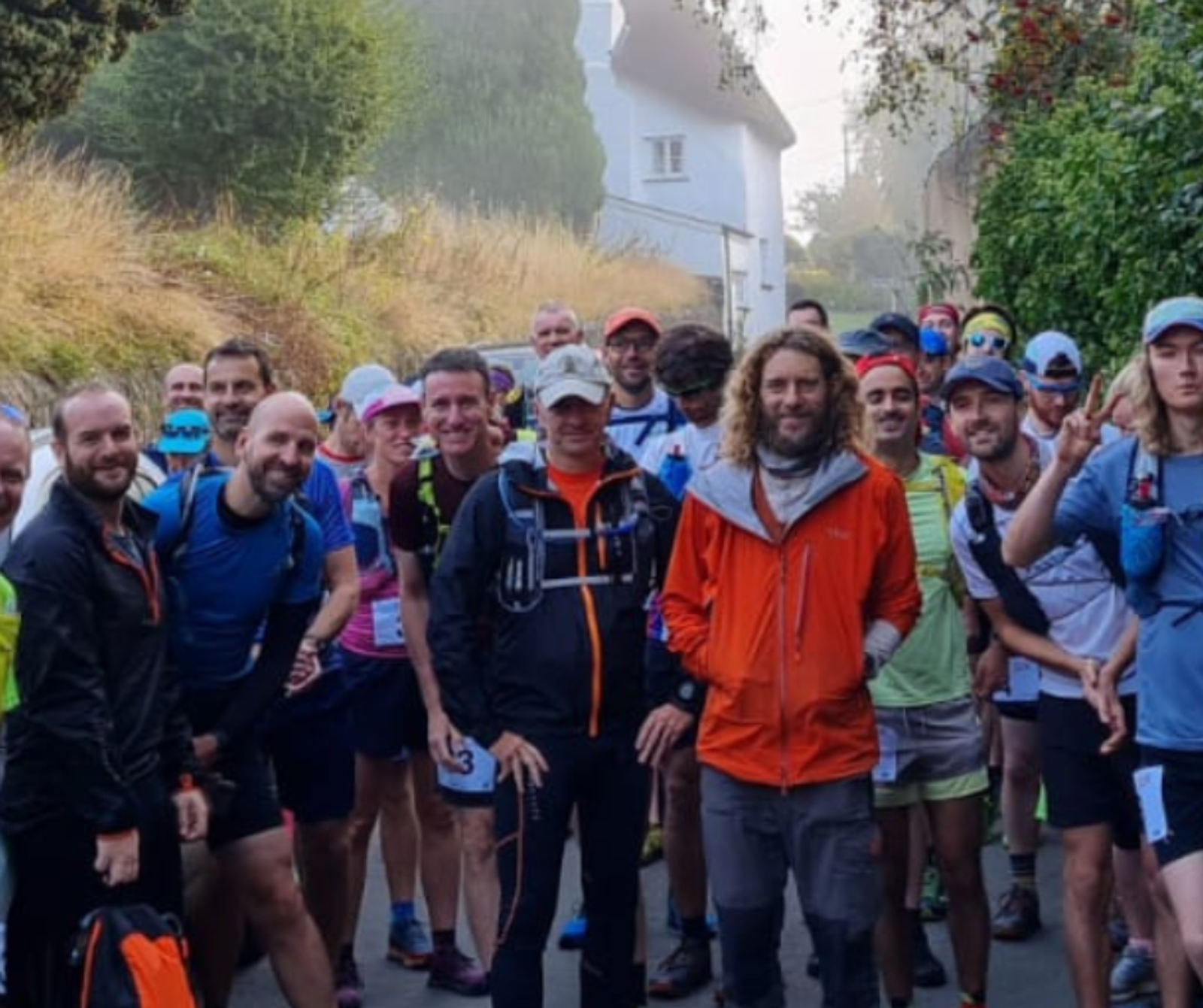 A group of runners gathers at the starting line of an outdoor race. They wear various athletic clothing and gear, with some in bright colors and some in darker tones. A few runners smile and make peace signs at the camera, while others focus ahead. Houses and greenery are visible in the background.