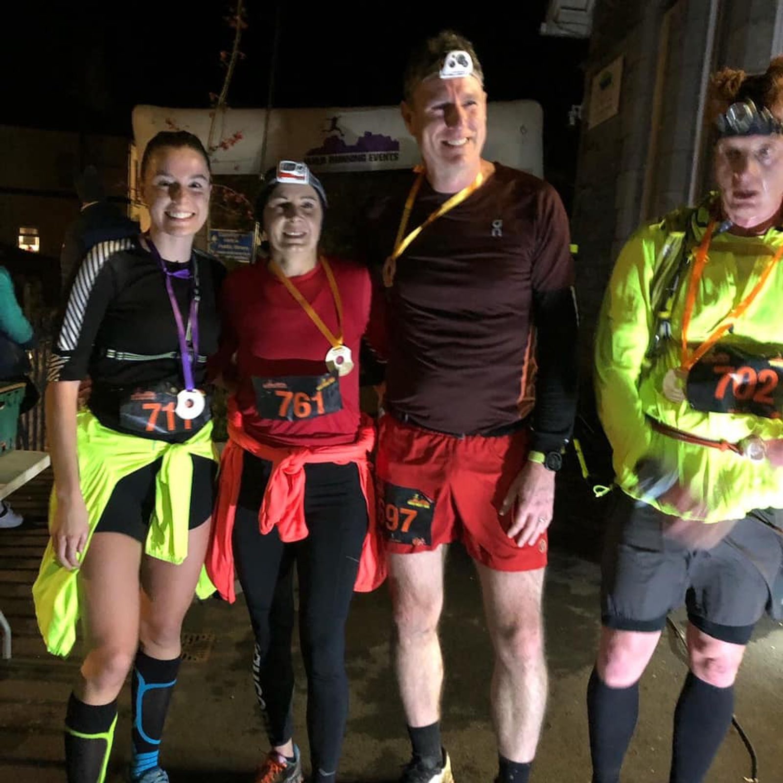 Four runners are standing together, smiling and wearing medals around their necks in a nighttime setting. They are dressed in athletic gear and have headlamps. Their race numbers, starting with 71, 761, 97, and 702, are prominently displayed on their outfits.