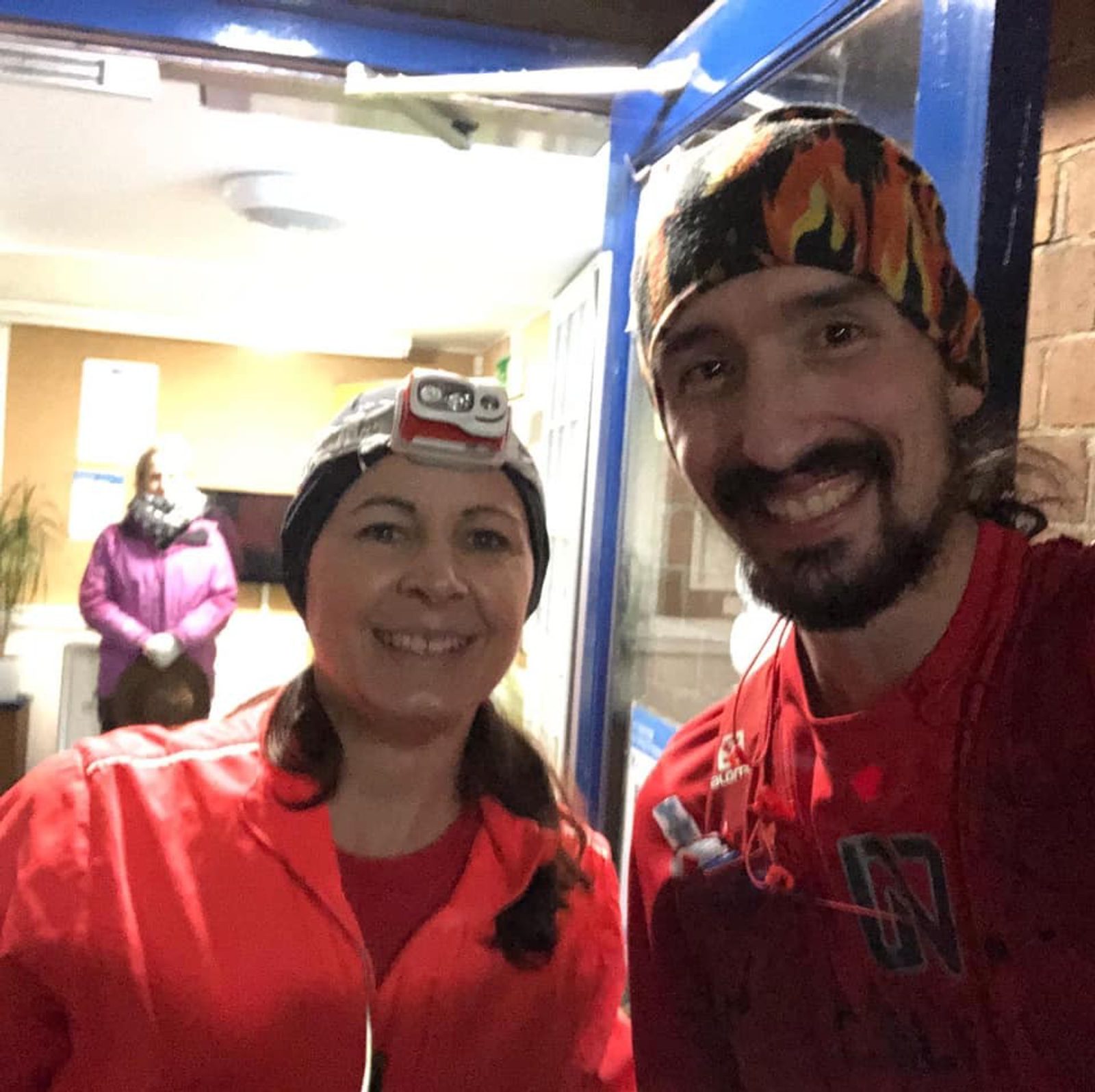 Two people in outdoor running gear stand smiling in front of a building entrance at night. Both are wearing headlamps and colorful headbands; the woman has a red and white headlamp, and the man has a flame-patterned headband. A person in a purple coat stands in the background.