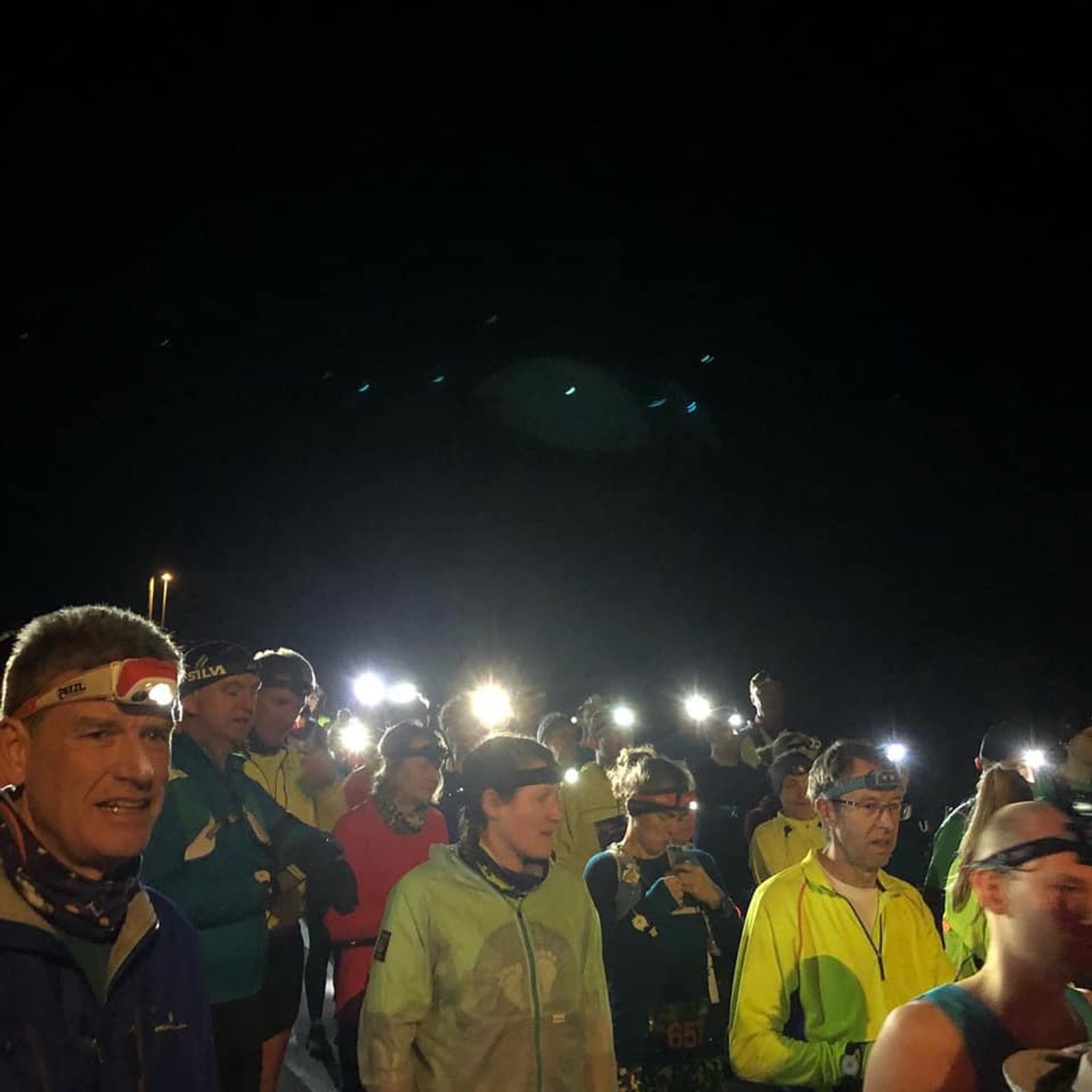 A group of runners, dressed in athletic gear and headlamps, are gathered in low-light conditions, likely before the start of a night race. Some are chatting and adjusting their equipment. The scene is illuminated by the runners' headlamps and a few distant lights.
