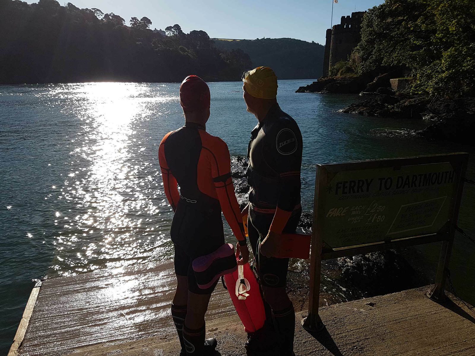 Two people in wetsuits and swim caps stand on a ramp near the edge of a river, holding swim floats. The sun is low, casting a bright reflection on the water. A sign pointing towards a ferry to Dartmouth is visible. Trees and a building are in the background.