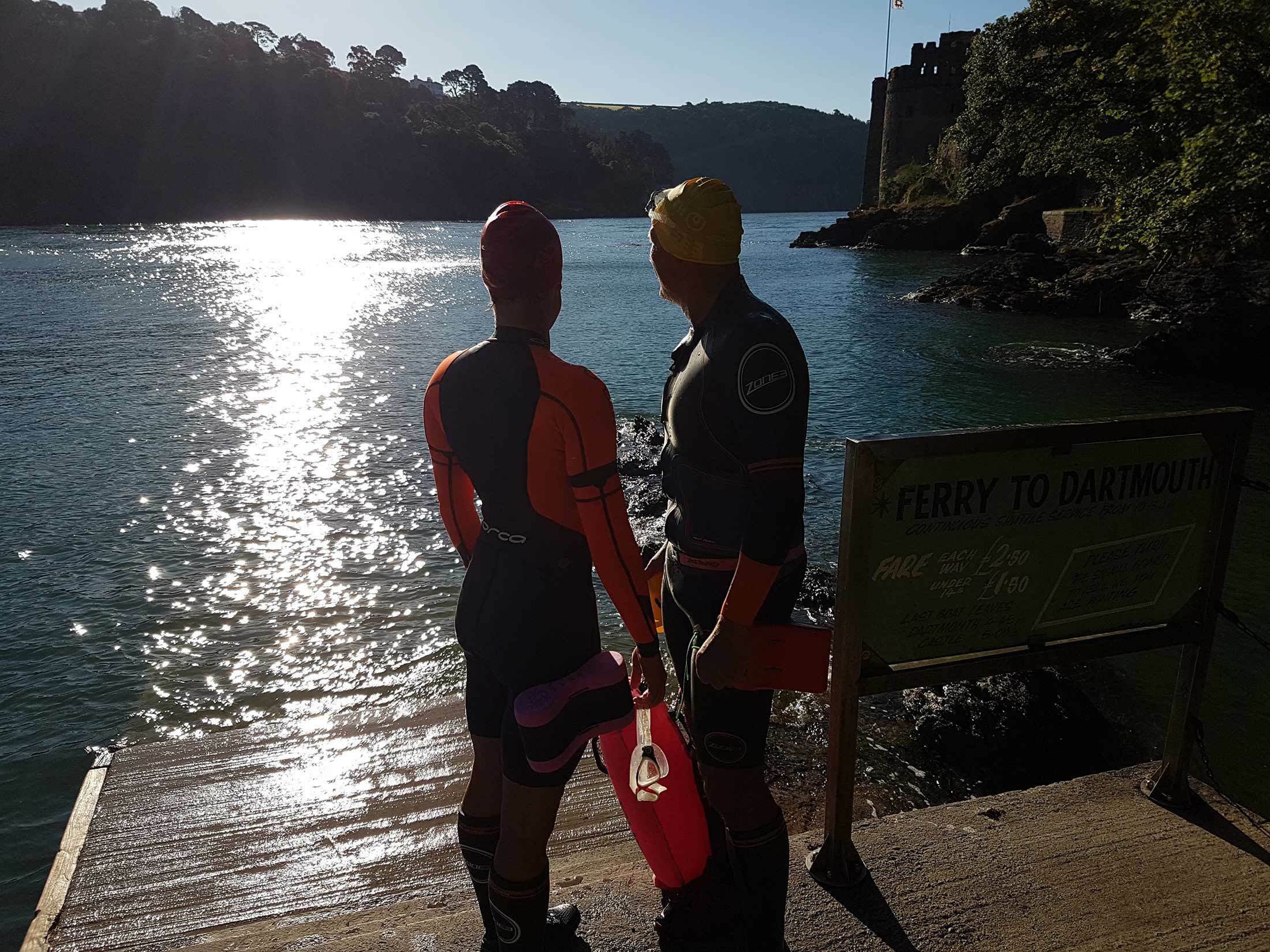 Two people in wetsuits and swim caps stand on a ramp near the edge of a river, holding swim floats. The sun is low, casting a bright reflection on the water. A sign pointing towards a ferry to Dartmouth is visible. Trees and a building are in the background.