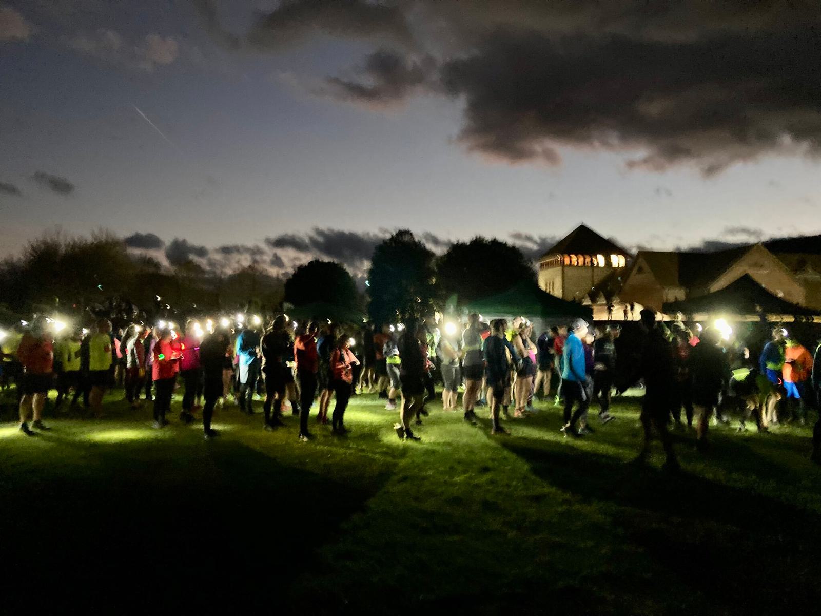 A group of people gathered outside at dusk, illuminated by headlamps and flashlights. The sky is dark, with clouds visible. A large building with lights glowing from its windows stands in the background, partially silhouetted against the deepening sky.