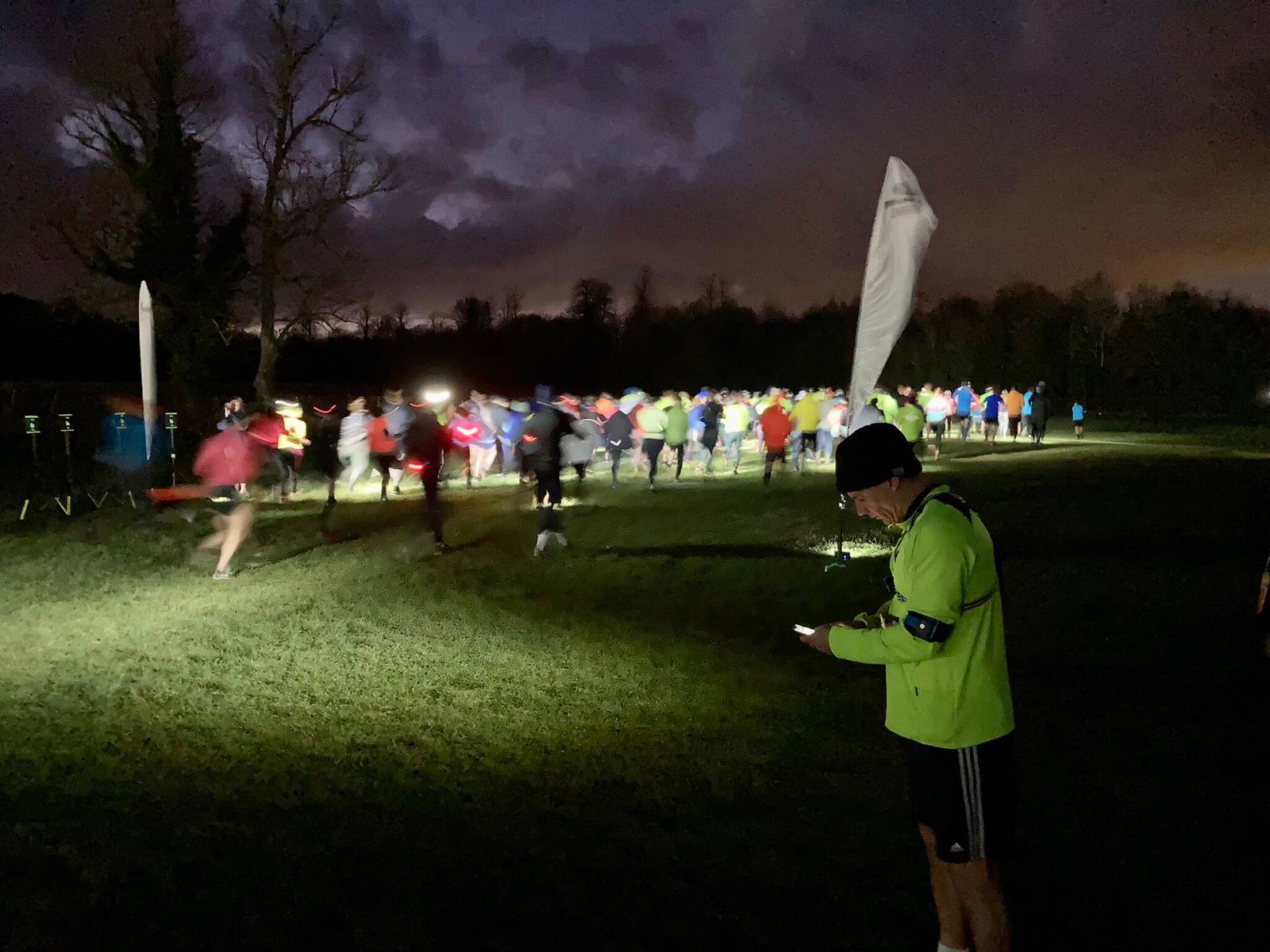 A group of runners, wearing brightly colored athletic gear, are participating in a nighttime race on a grassy field. The darkness is lit by scattered lights, and a person in a green shirt stands in the foreground, checking a device. Trees line the background.