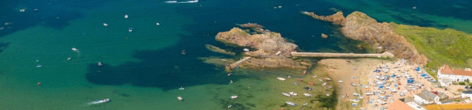 Aerial view of a coastal area with a rocky pier extending into the sea. Boats dot the clear, turquoise water. A sandy beach with people and buildings is visible on the right. The scene includes lush greenery on the rocky terrain.
