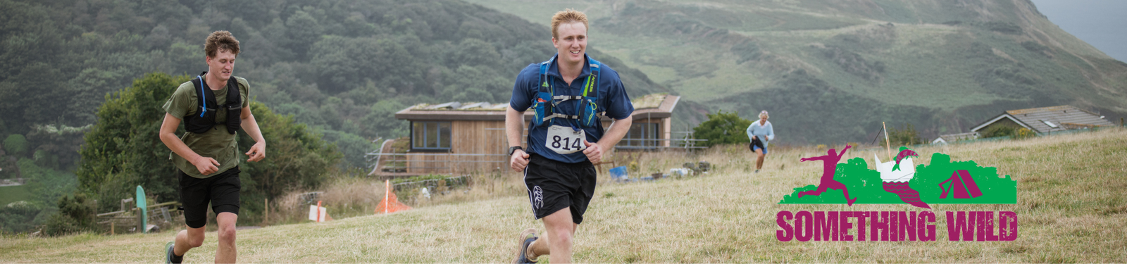 Two runners are competing in an outdoor race, wearing running gear with bib numbers. They are on a grassy trail with hills and houses in the background. A "Something Wild" logo with running figures and a tent is in the foreground.