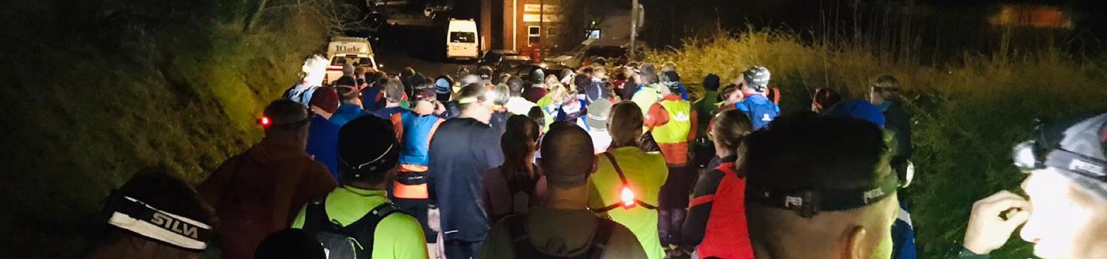 A large group of people wearing headlamps participate in a nighttime event on a road surrounded by grass and trees. They are dressed in athletic gear and jackets, walking or running toward a lit area in the distance.