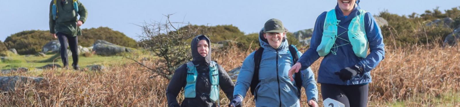 A group of four people are trail running on a grassy, rocky path surrounded by shrubs. They are wearing outdoor gear, including backpacks and bright running vests. The sky is clear and sunny.