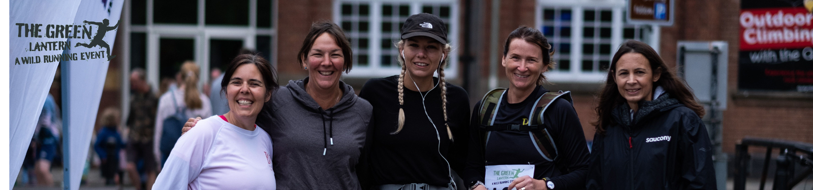 Five people standing together outdoors, smiling at the camera. They are dressed in athletic gear. A banner to the left reads "The Green Lantern Wind Running Event." A building is visible in the background.
