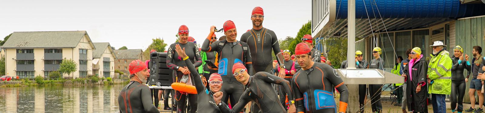 A group of people in wetsuits and red swim caps prepare for a swimming event by a lakeside. Some are smiling and posing for the camera, while others adjust their gear. Spectators and swimmers in bright green jackets stand nearby.