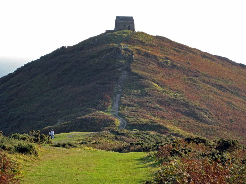 A small stone chapel sits atop a grassy hill with a winding path leading up to it. Surrounding the hill are patches of green, brown, and orange vegetation. Two people are seen walking at the base of the hill under a clear sky.