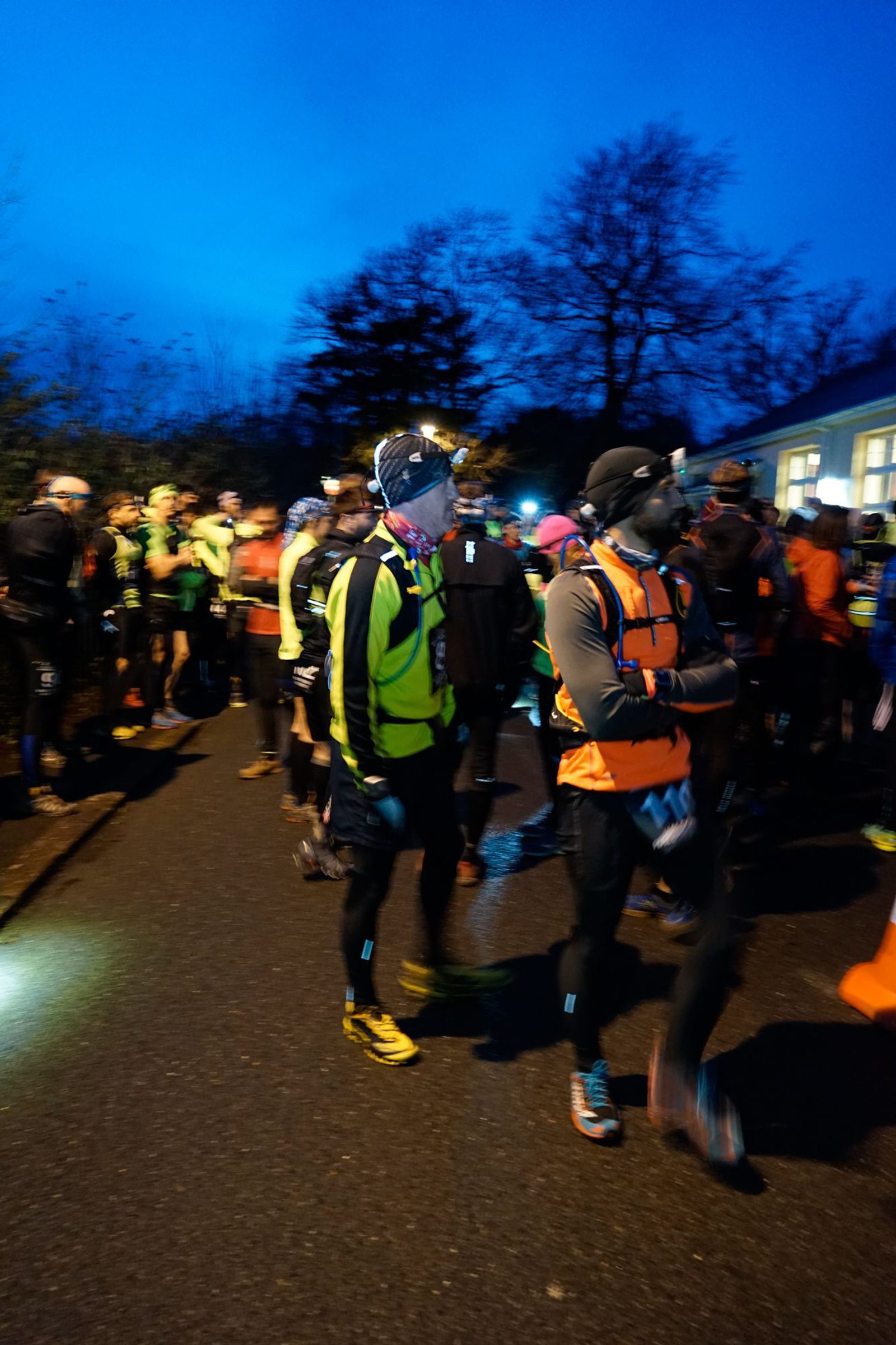 A group of runners in athletic gear gathers on a road at dusk, preparing for a race. Some are wearing headlamps and reflective gear. The scene is dimly lit, creating a sense of early morning or evening. Trees and a building are visible in the background.