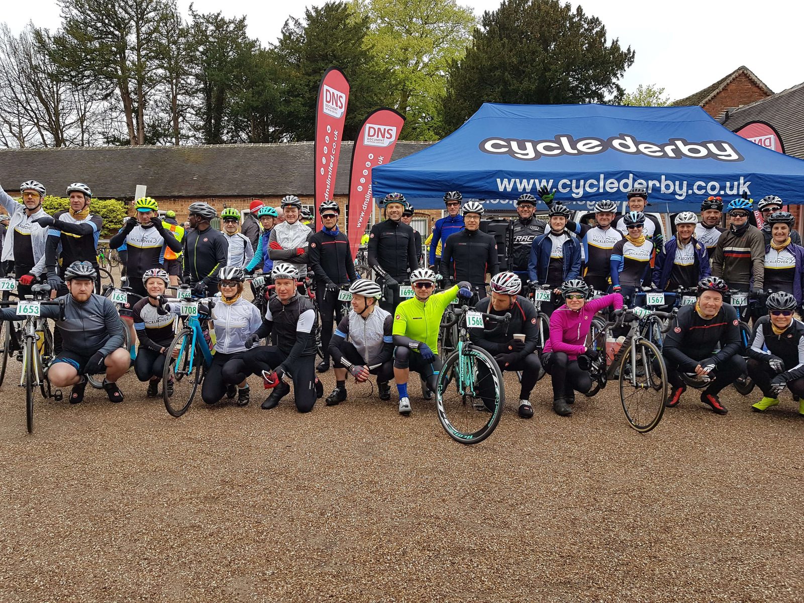 A large group of cyclists poses in front of a "Cycle Derby" tent. They are dressed in cycling gear, helmets, and sunglasses, standing with their bicycles on a gravel surface. The background features several trees and red banners with "DNS" text.