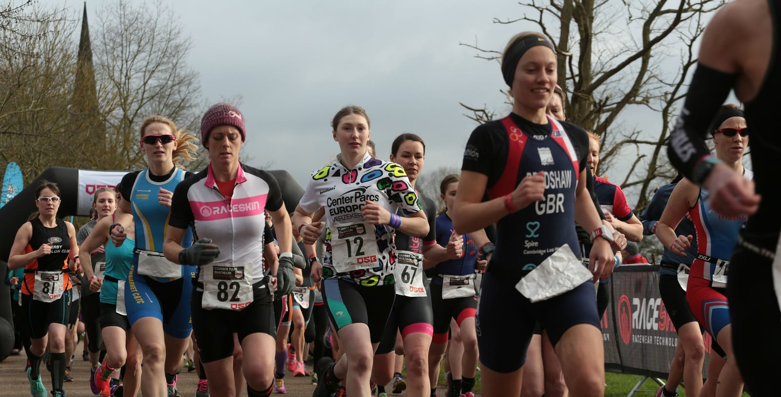 A group of athletes run in an outdoor race. They wear sport attire and race bibs with numbers visible on their clothing. Bare trees and a cloudy sky are in the background. Participants appear focused and determined. A race event banner is seen to the right.