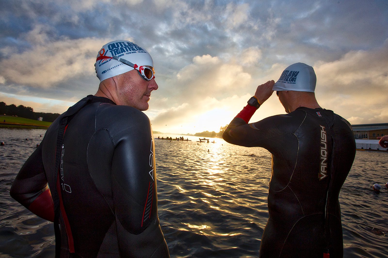 Two athletes in wetsuits and swim caps prepare for a triathlon swim at dawn by a lake. One athlete adjusts his cap while the other looks onward. The sky is partly cloudy with the sun rising, casting a warm glow over the water.