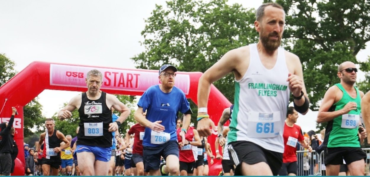 A group of runners, wearing various athletic gear, begin their race under a large red "START" arch. The foreground shows a man in a white tank top numbered 661, and beside him, men in blue, black, and green shirts run forward. Trees are visible in the background.