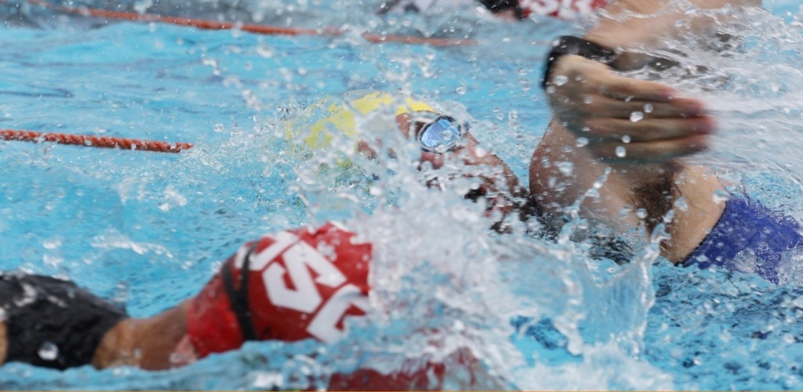 A swimmer wearing goggles and a cap emerges from the water during a race, with splashes of water surrounding them. The swimmer is mid-stroke, and another swimmer's arm is visible in the background. The scene is dynamic, capturing the intensity of the competition.