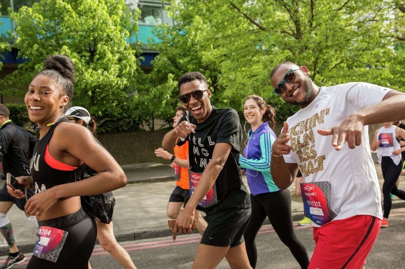 A group of runners participating in an outdoor race, smiling and energetic. They are dressed in athletic wear with race bibs, and the background shows a tree-lined street and greenery. It’s a sunny day, and everyone appears to be enjoying the event.