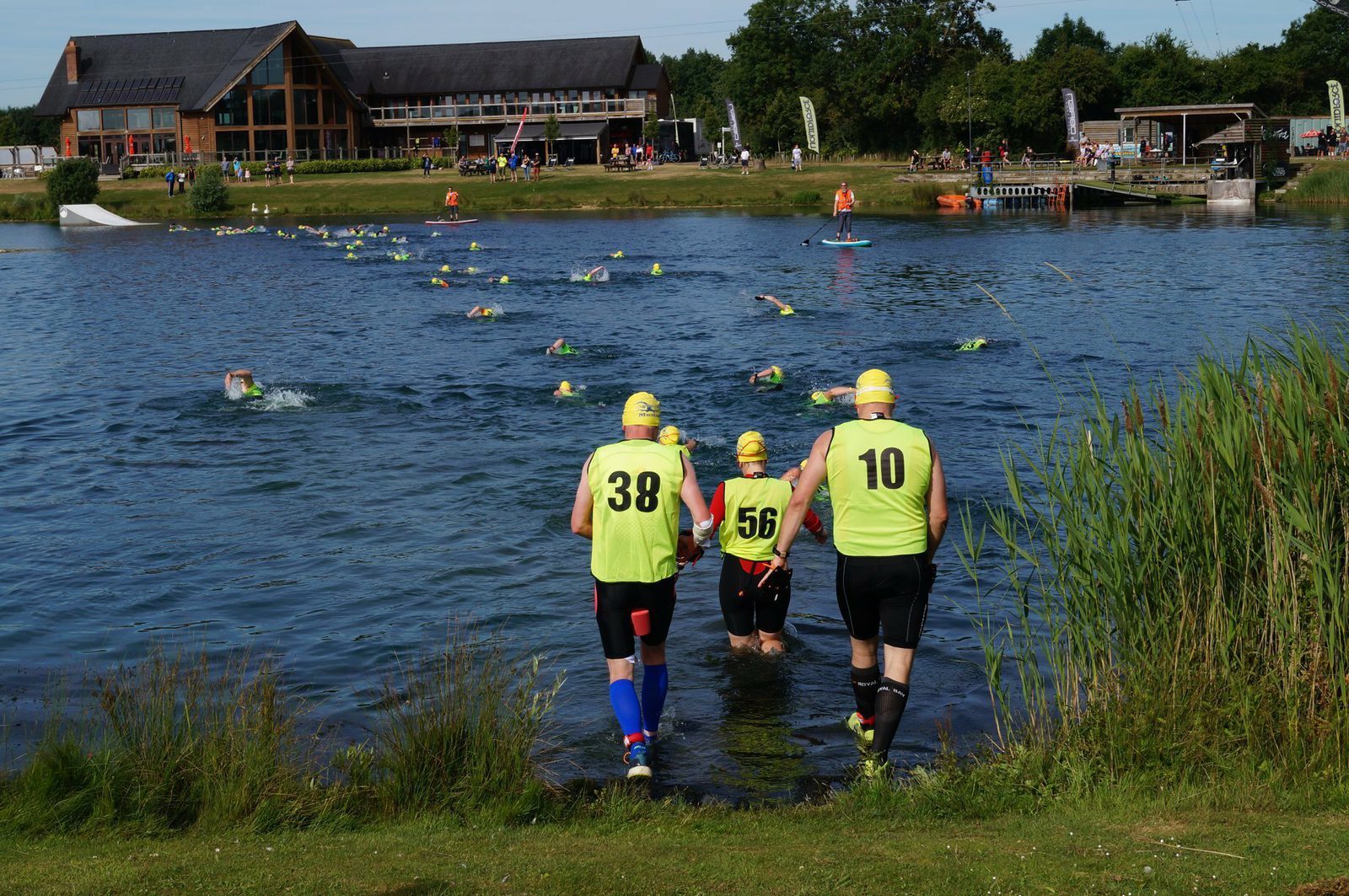 Three athletes in high-visibility vests, numbered 38, 56, and 10, are entering a lake for a swim. More swimmers with yellow swim caps are ahead in the water. A building and spectators can be seen in the background. The event seems to take place on a sunny day.