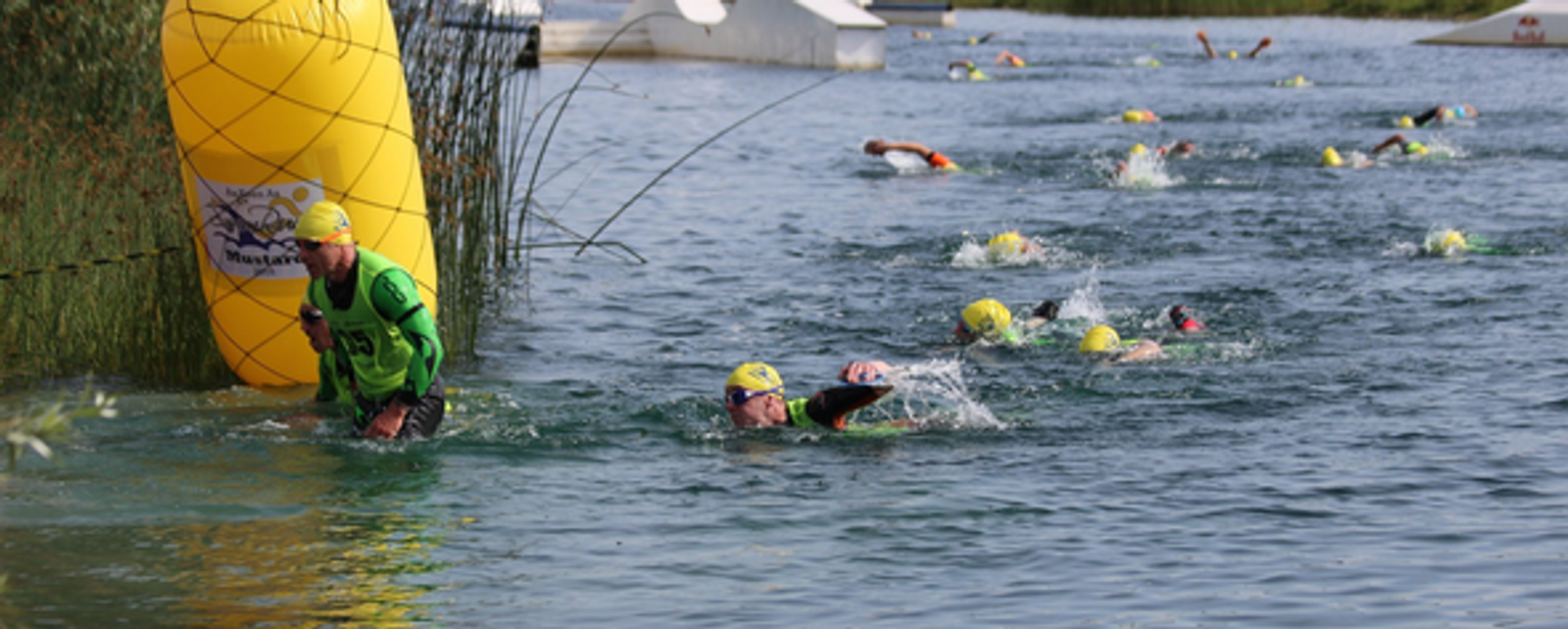 Triathletes wearing swim caps and wetsuits swim through a narrow waterway, guided by a large yellow buoy. A man in a green outfit holding a megaphone stands in shallow water, waving a pointer. Participants can be seen in different stages of the swim.