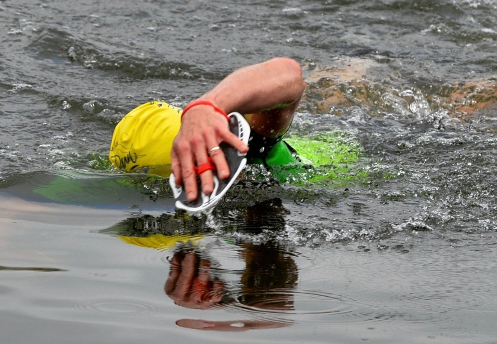 A swimmer in a green wetsuit and yellow swim cap competes in an open water event, with one arm extended forward and wearing a white hand paddle. The water ripples around them as they stroke through it. They also have a red band on their wrist.