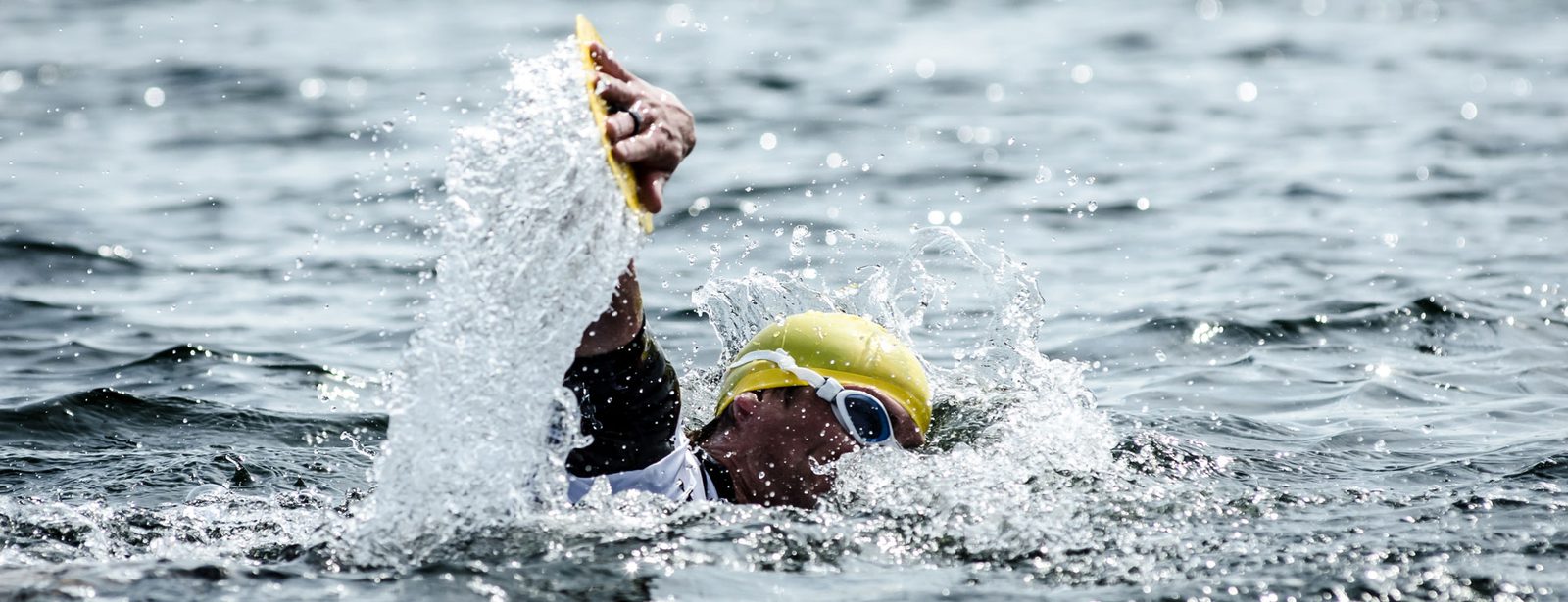 A swimmer wearing a yellow swim cap and goggles powers through the water during an open-water swim. The swimmer's arm is raised mid-stroke, splashing water, and creating dynamic movement. The water's surface is slightly rippled with light reflections.