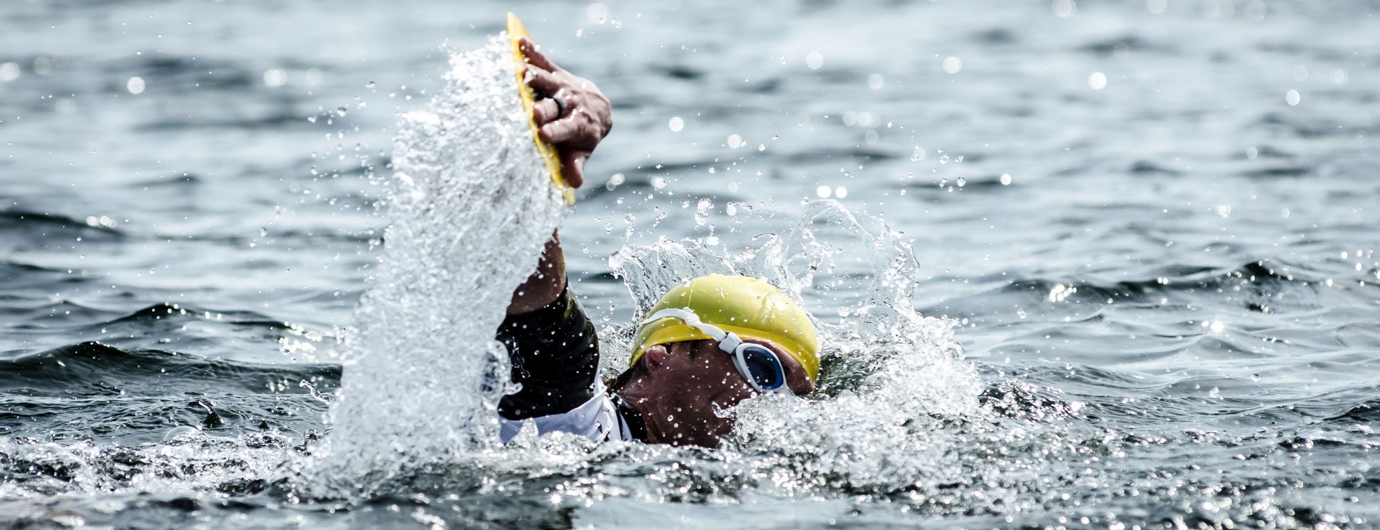 A swimmer wearing a yellow swim cap and goggles powers through the water during an open-water swim. The swimmer's arm is raised mid-stroke, splashing water, and creating dynamic movement. The water's surface is slightly rippled with light reflections.