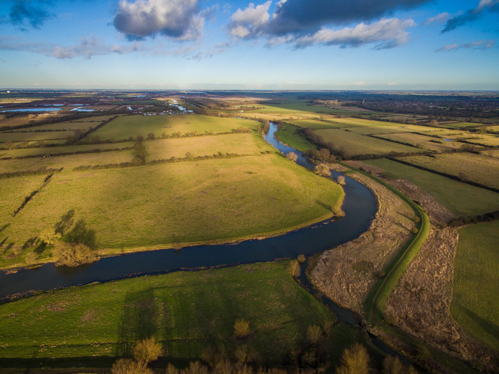 Aerial view of a serene landscape with a winding river cutting through green fields under a blue sky with scattered clouds. The river meanders through the countryside, bordered by patches of trees and bushes, casting shadows in the late afternoon sun.