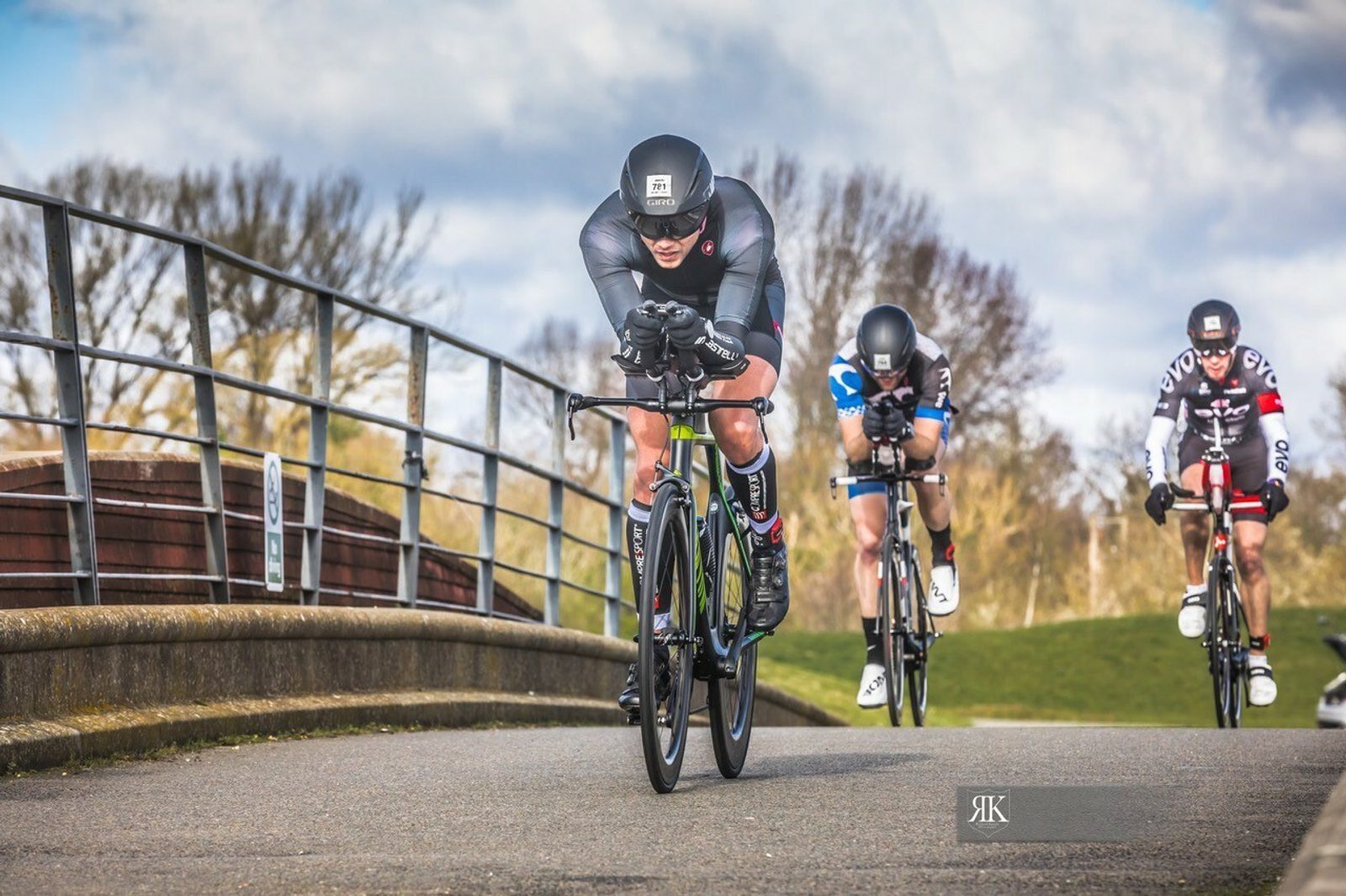 Three cyclists in racing gear and helmets are competing on a paved road with metal railings on the left side and a grassy area to the right. The leading cyclist is in a full aero position, with two cyclists closely following. The weather is clear with scattered clouds.