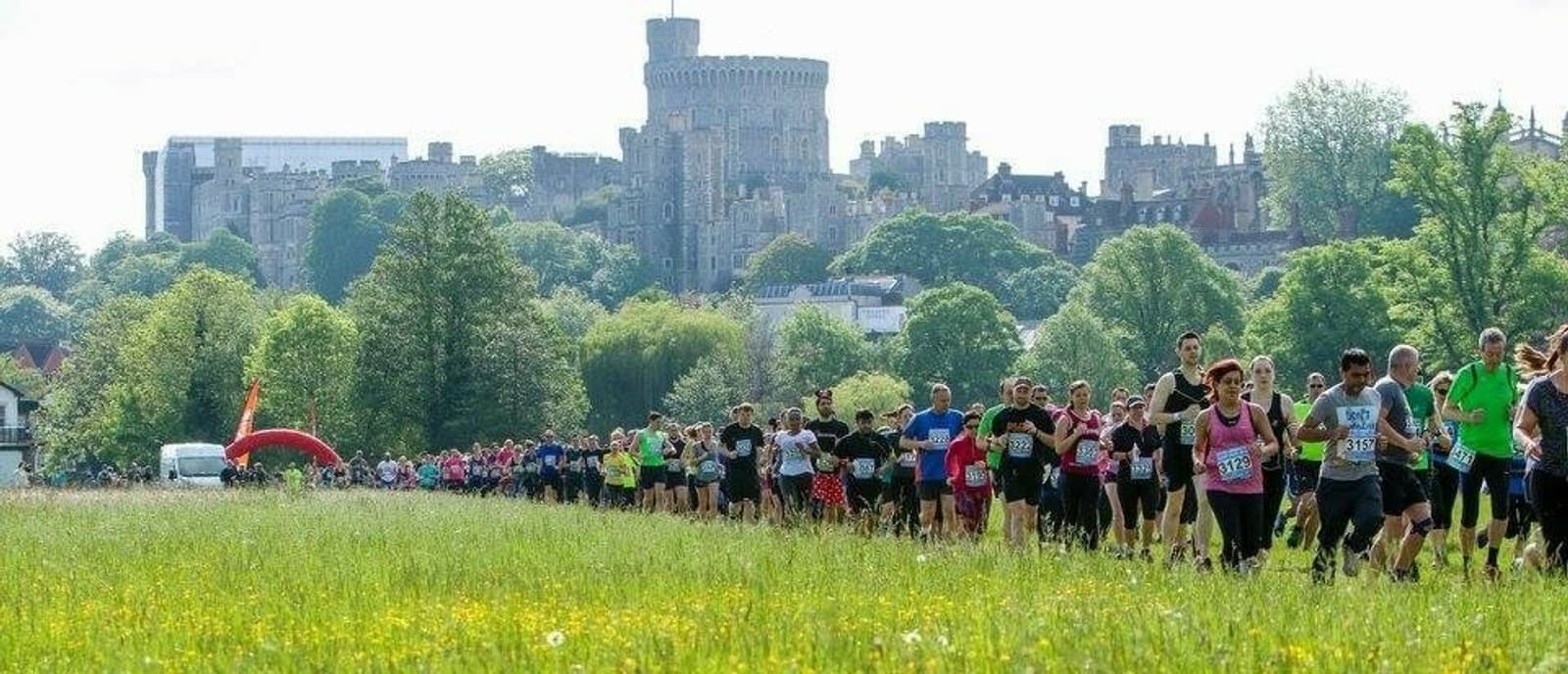 A large group of runners, dressed in colorful athletic attire, participate in a race through a lush, green field with a historic castle and tall trees in the background. The sky is clear, and the atmosphere appears energetic and vibrant.