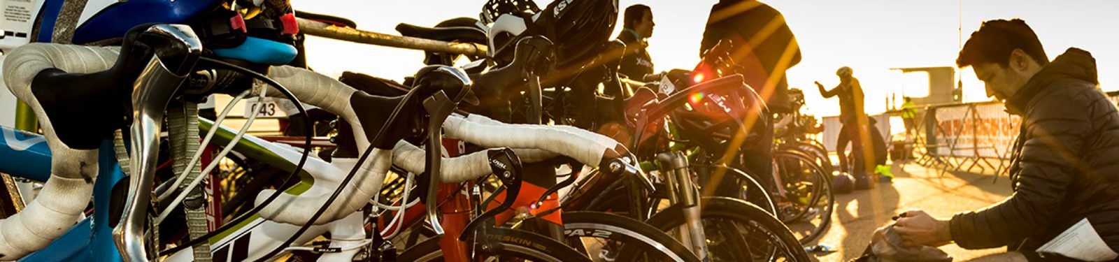 Several bicycles are lined up at a transition area during a triathlon, with their handlebars wrapped in white tape. Participants can be seen preparing in the background as the sun rises, casting a warm glow over the scene. A man in a jacket adjusts his shoes in the foreground.