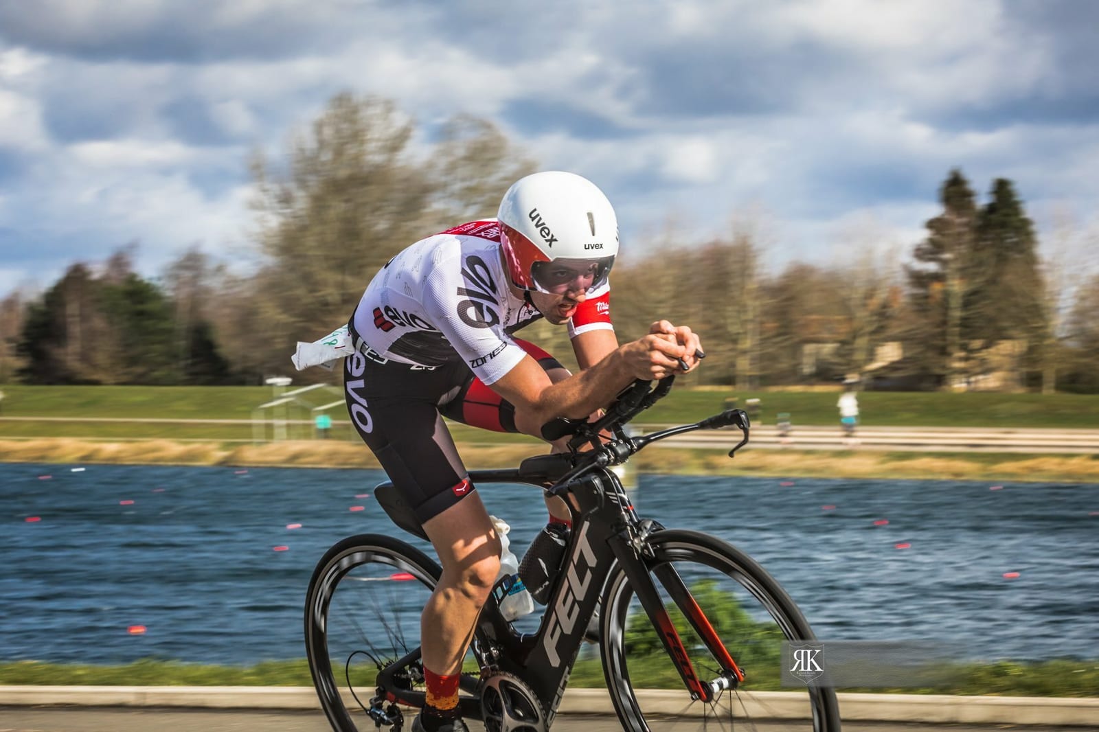 A cyclist wearing a white helmet and aerodynamic cycling gear rides a black bicycle on a road beside a body of water, with a grassy area and trees in the background under a partly cloudy sky.