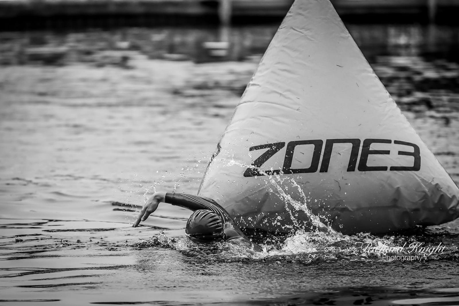 A swimmer in a wetsuit and swim cap is swimming in open water, turning around a large triangular buoy labeled "ZONE3." The water is splashing as the swimmer's arm is outstretched, making a stroke in the water.