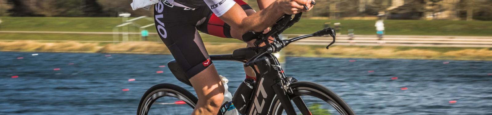 A cyclist wearing a white and black outfit rides an aerodynamic bike along a waterfront path. The background features a body of water with scattered buoys, grassy areas, and blurred figures of other people walking or cycling in the distance.