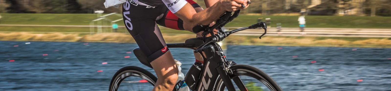 A cyclist in full gear rides a racing bike along a waterfront path. The cyclist is wearing a white and red jersey, black and red shorts, a helmet, and sunglasses. In the background, there is a body of water and a blurred figure jogging.