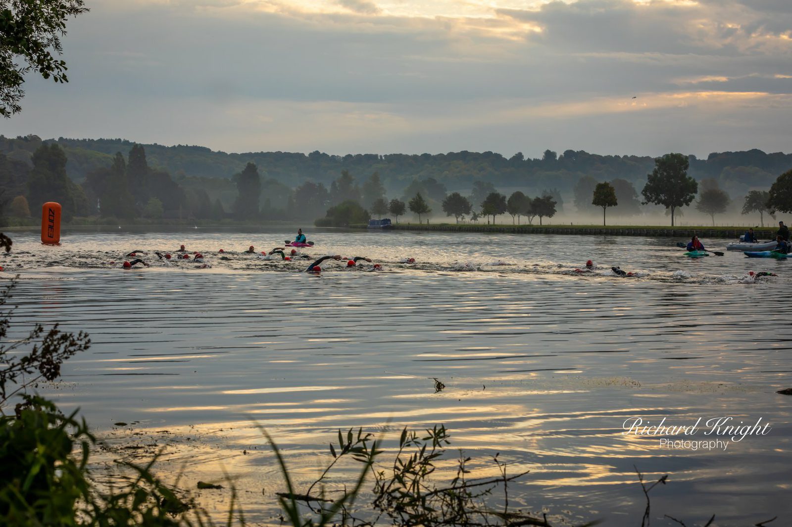 A group of swimmers in a misty lake swim towards an orange buoy during dawn. Silhouetted trees and hills form the backdrop under a cloudy sky with streaks of sunlight. Vegetation is visible in the foreground, and kayakers accompany the swimmers for safety.