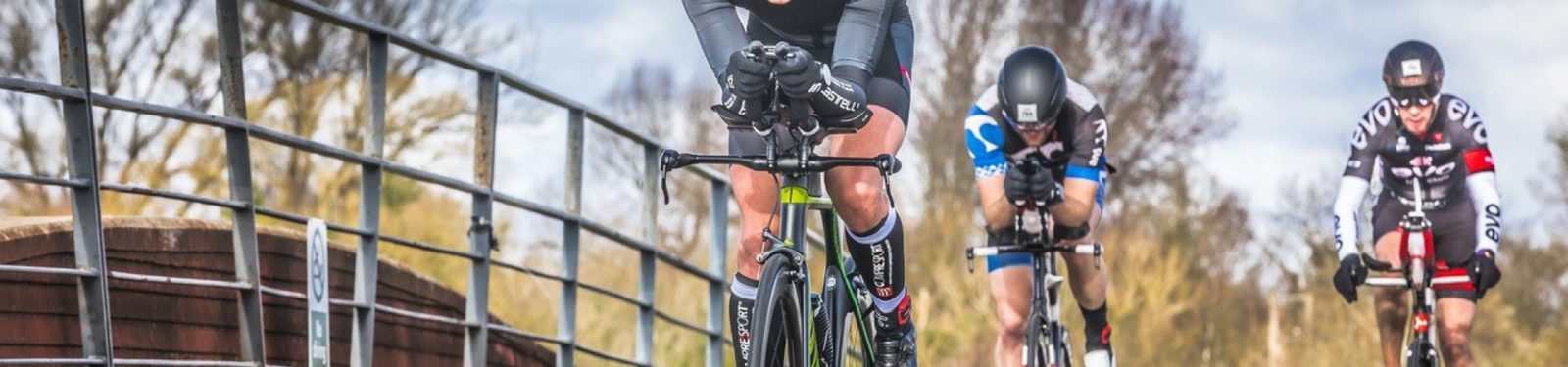A group of three cyclists in vibrant cycling attire riding on a paved road. The background features trees and a cloudy sky. The cyclists appear focused, with the lead rider in the foreground, followed by two others close behind. A metal fence runs alongside the road.