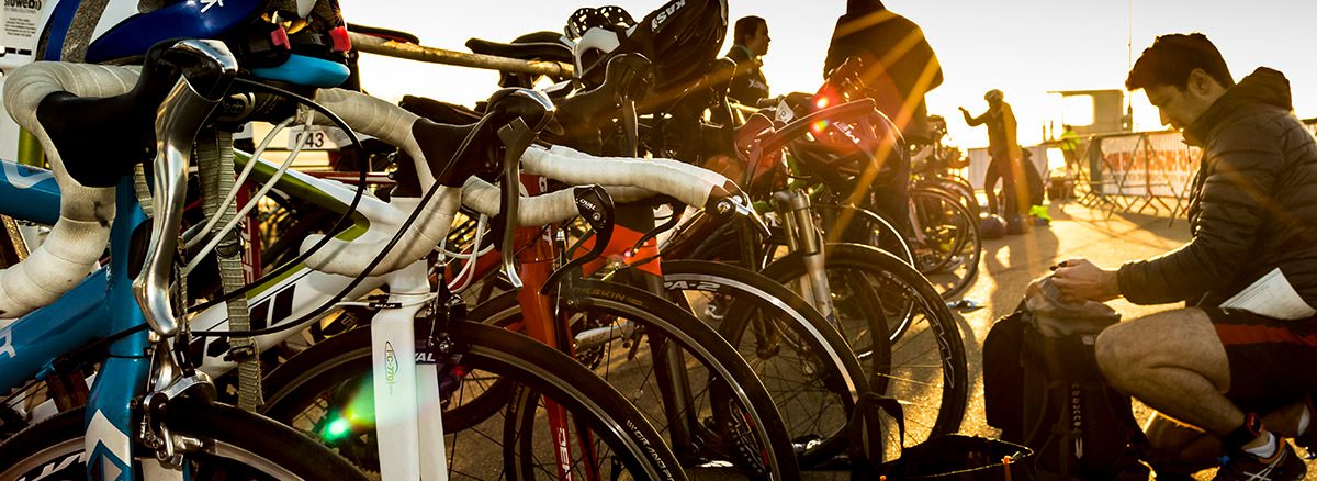 A row of road bikes are parked on a bike rack, with cyclists preparing their gear in the background as sunlight shines brightly. The scene is likely at a race or training event, with people dressed in cycling gear and focusing on their equipment.