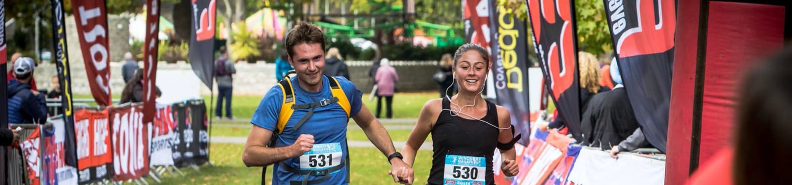 Two runners are crossing the finish line in a park setting. Both wear numbered bibs, with the man's reading 531 and the woman's 530. The woman is smiling and wearing earphones. Vibrant banners line the path and other people are visible in the background.