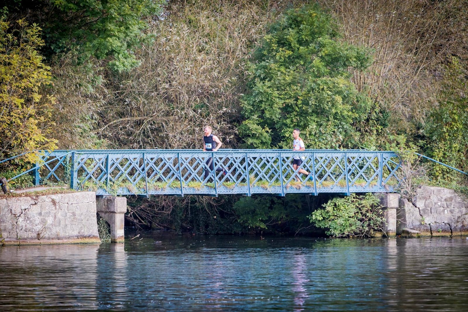 Two people are jogging across a blue metal bridge over a calm river. The background features dense green foliage and trees, and the bridge's stone supports are visible. The scene is peaceful and scenic.