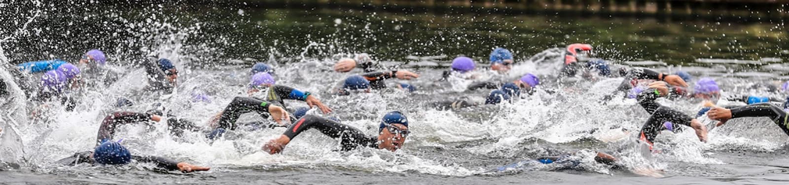 A group of swimmers in wetsuits and swim caps are actively swimming in open water, creating a large splash. The image captures the intensity and movement of multiple athletes competing in what appears to be a triathlon or open water race.