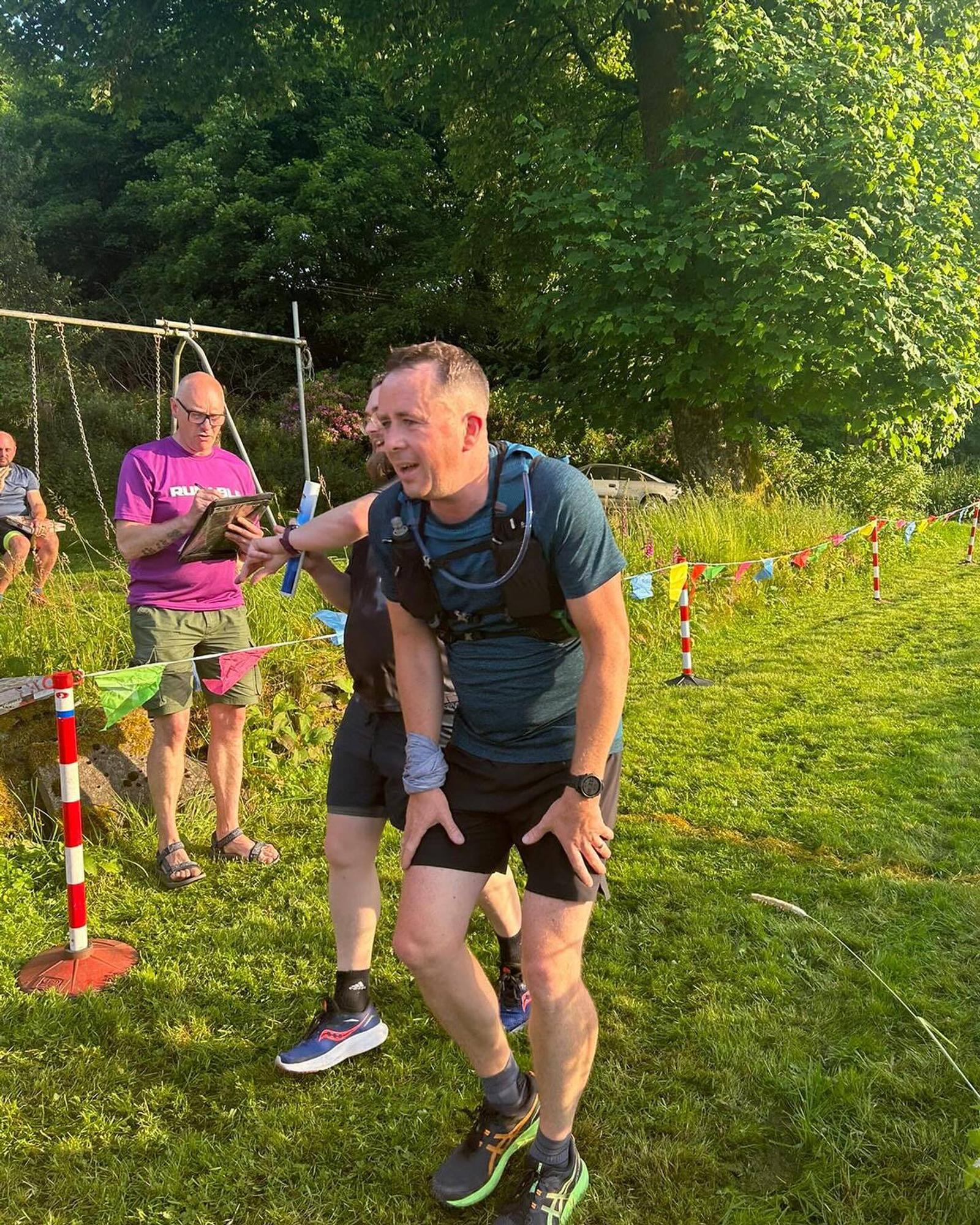 A man in a blue shirt and black shorts appears exhausted while participating in an outdoor event. A person in a purple top is checking something on a clipboard behind him. They stand on grassy ground with leafy trees in the background.