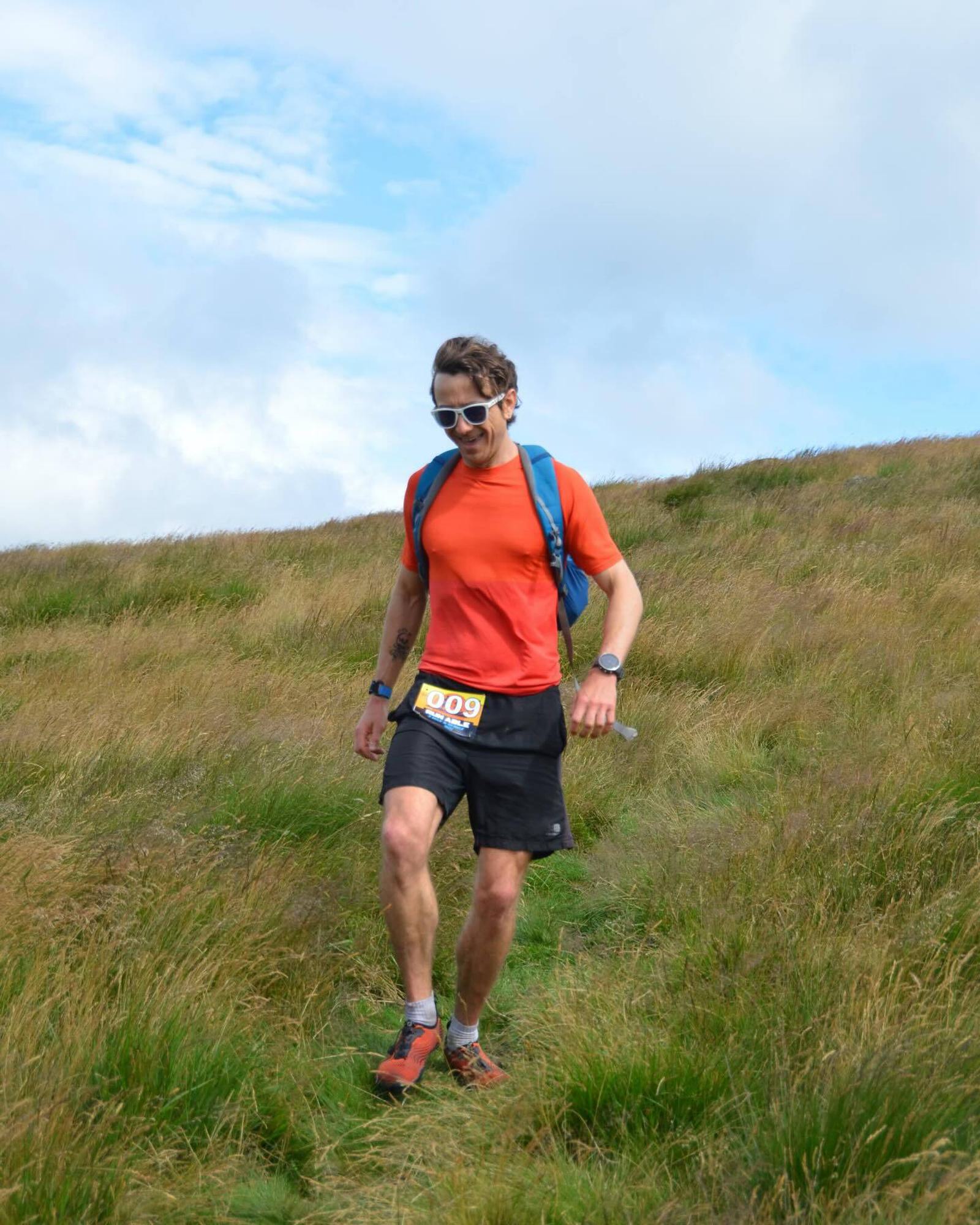 A person in an orange shirt and black shorts is running down a grassy hill, wearing sunglasses and a blue backpack. The runner has a race number pinned to their shorts. The sky is partly cloudy.