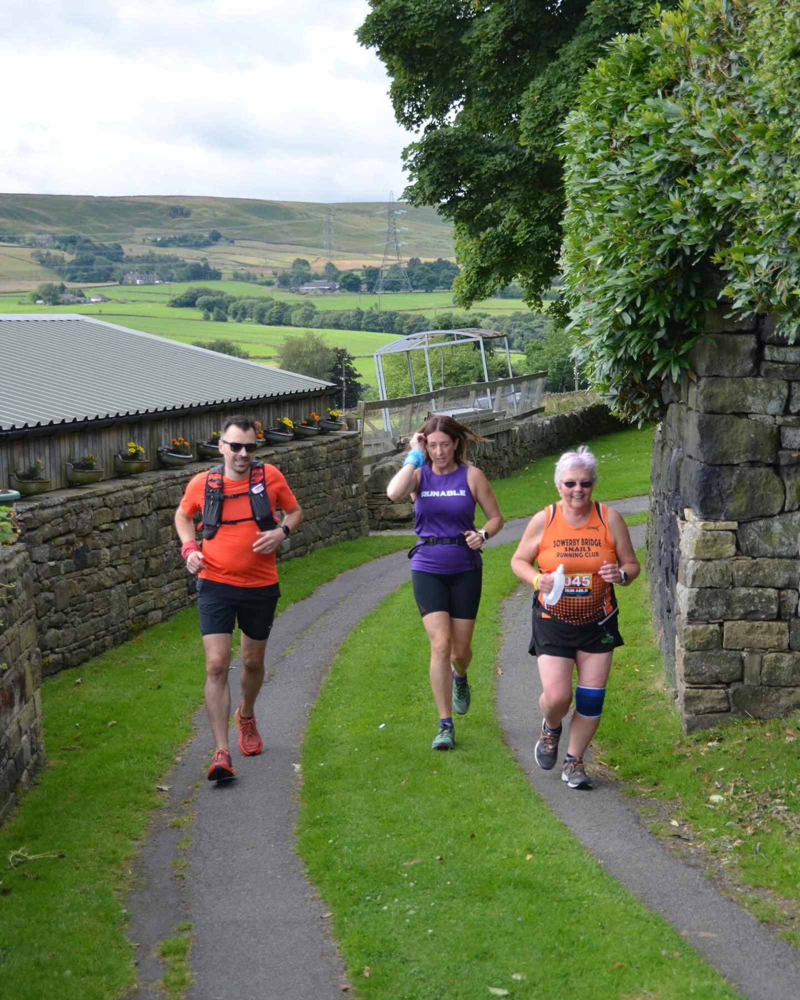 Three people are jogging along a narrow path between stone walls in a rural area. They wear colorful athletic gear, and one has a race bib. The background features lush greenery and hills under a partly cloudy sky.