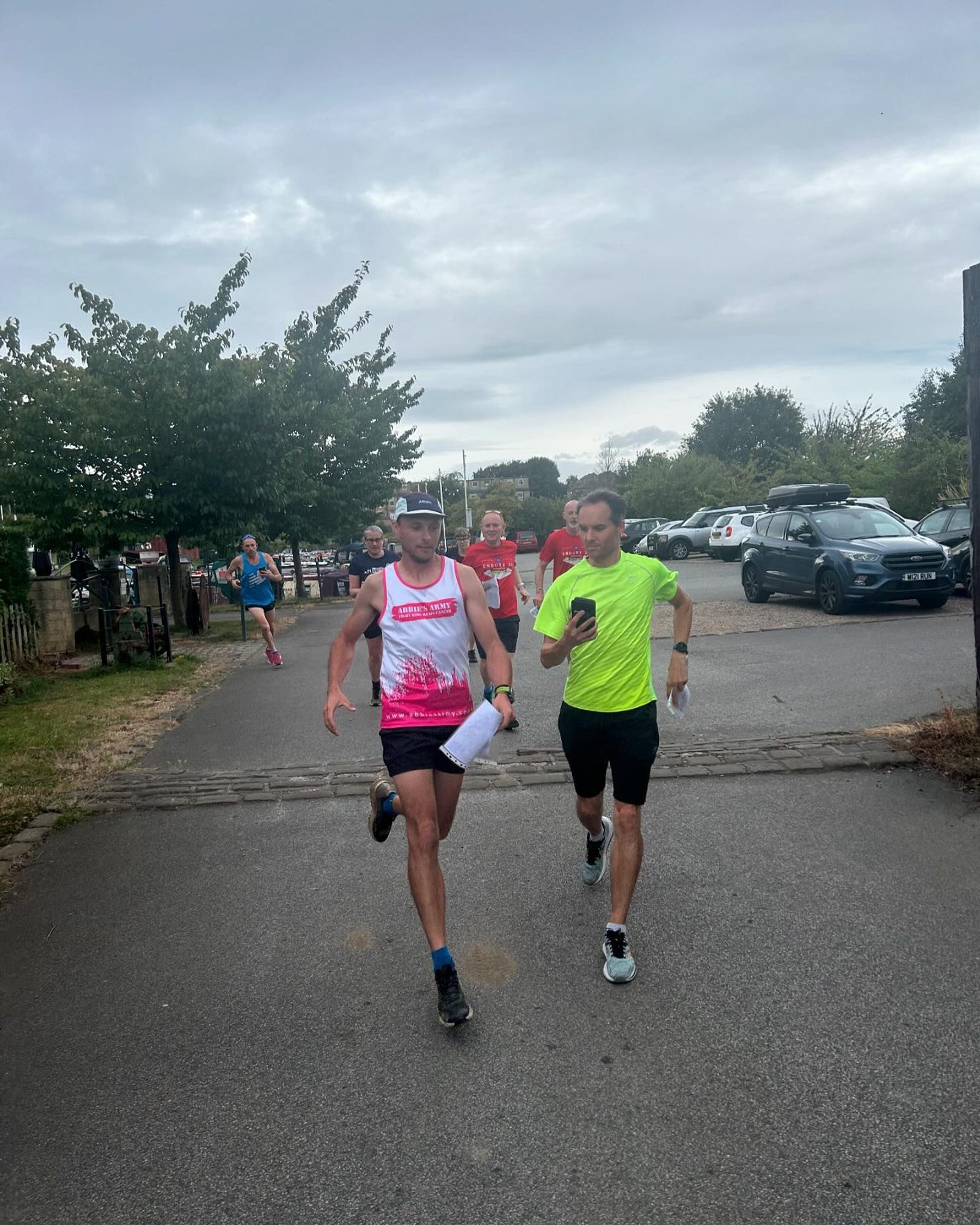 Two men are running on a paved path; one in a white and pink tank top and the other in a neon yellow shirt. They are near parked cars and trees, with a few other runners in the background under an overcast sky.
