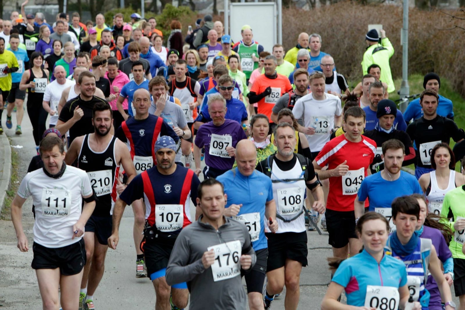 A large group of runners participate in a road race, wearing various athletic gear and bib numbers. Some smile and others concentrate on their run, demonstrating a range of emotions and energy levels. Trees and spectators are visible in the background.