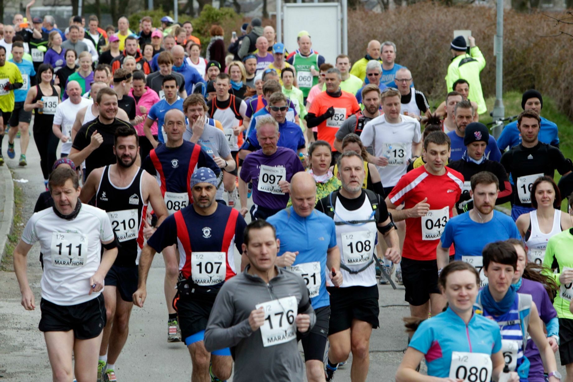 A large group of runners participate in a road race, wearing various athletic gear and bib numbers. Some smile and others concentrate on their run, demonstrating a range of emotions and energy levels. Trees and spectators are visible in the background.