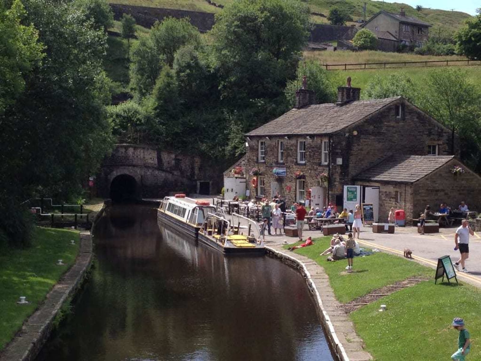 Scenic view of a narrow canal with a long boat docked near a quaint stone building. People are sitting at outdoor tables and walking along the canal's edge. Lush greenery surrounds the area, and a tunnel is visible in the background.